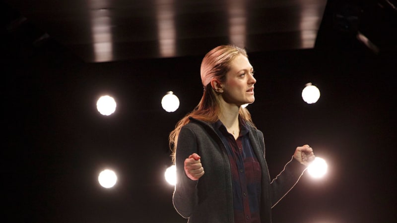 A blonde woman wearing a flannel under a zip up hoodie and jeans stands in front of a square panel with sixteen circular lights backlighting her.