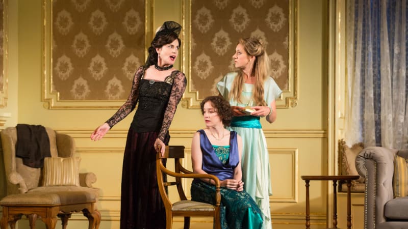 Three woman in 1910s party dresses. One wears a lacy black and burgundy dress with a fancy head piece. The other two wear blue and green dresses, one sitting, one standing.