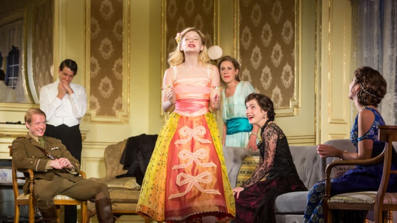 A young blonde woman in a bright peach and yellow dress with ribbons speaks passionately in a fancy 1910s living room while the people around her chuckle.