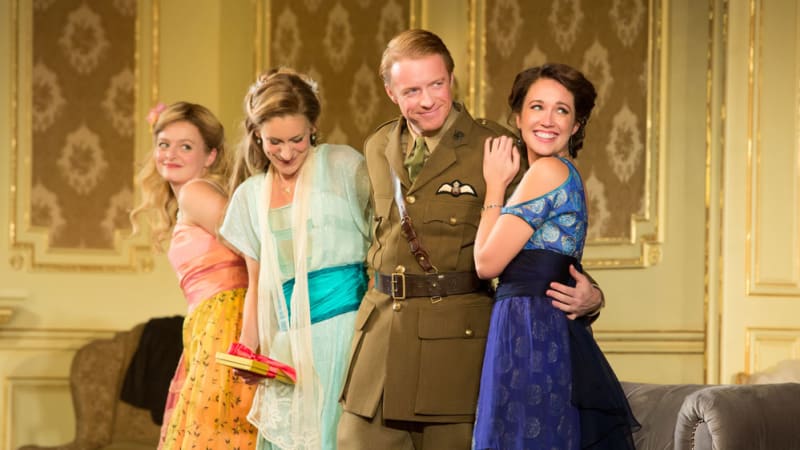 Three young woman in colorful 1910s party dresses cling bashfully to a handsome man in a military uniform.