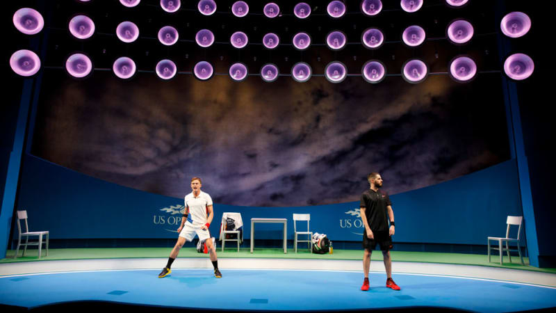 Two tennis players stand across from each other on a stage meant to look like a large, US Open tennis court.