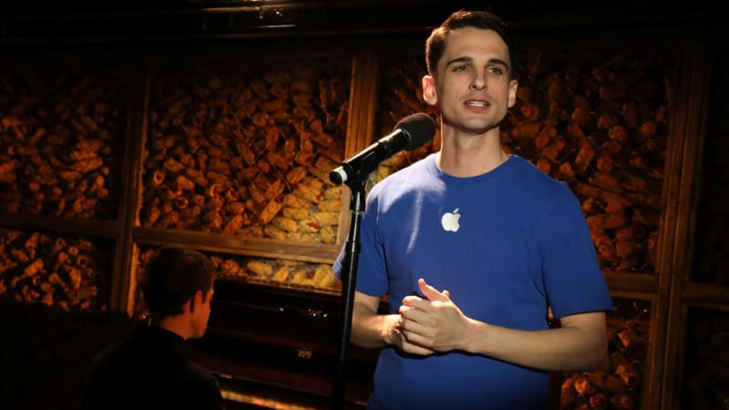 A man in a blue t-shirt with a Mac Apple logo on it sings at a microphone. Behind him, someone plays piano in front of a wall made of corn.