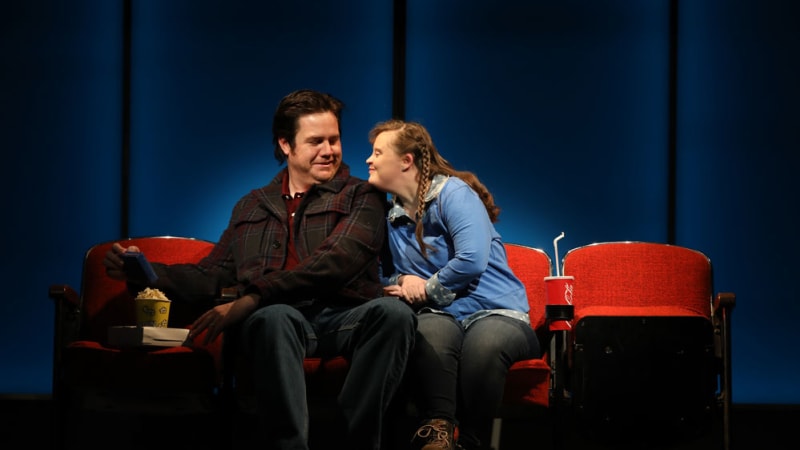 A man and woman on a date at a movie theater. The woman has Down syndrome and long brown hair. She rests her chin on the shoulder of the man, who smiles at her.