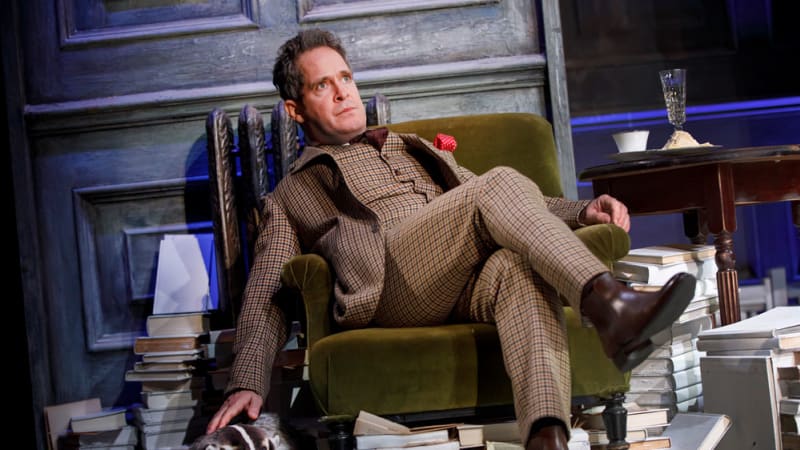 A man in a brown checkered suit set slumps in a velvet olive green chair with his legs crossed while his hand reaches down to pet a badger.