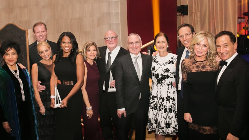 A group of people in formal attire line up for a photo together. Two of them hold awards.