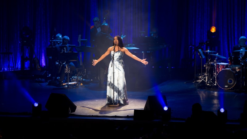 A woman sings on a blue-lit stage with her arms oustretched while a band plays behind her.