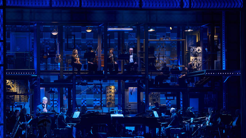 A stage with two levels of platforms contains a full orchestra. A man holds a microphone at a music stand to the left of the stage. Blue lighting washes over.