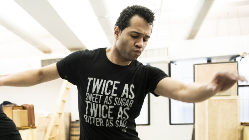 A man in a black shirt and pants dances smoothly in the rehearsal studio. 