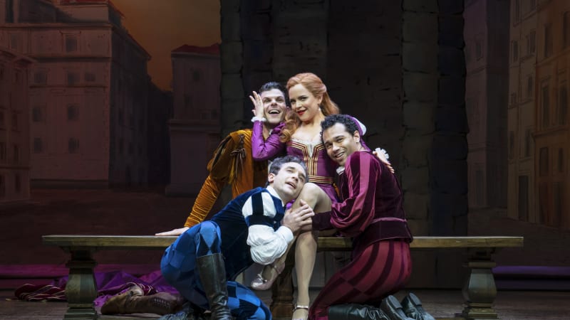A sultry woman with red hair in a short, purple, Shakespearean-styled dress sits on a bench while three men in Shakespearean garb dreamily hold and caress her.