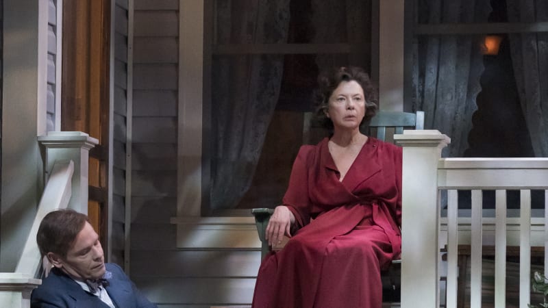 An older woman with brown curls and a red robe sits on a green rocking chair on the porch of a home. A man in a suit sits on the steps below her.