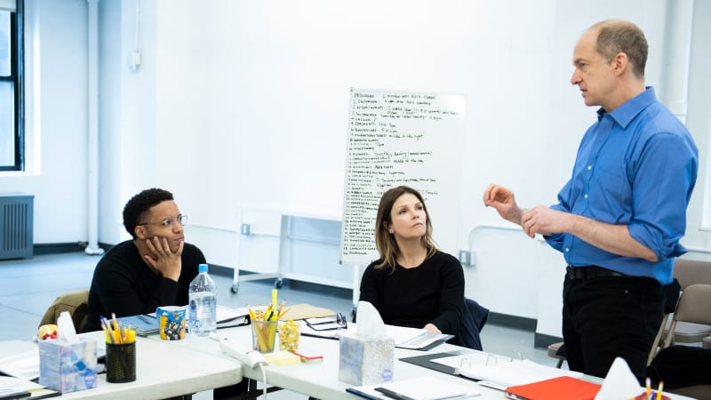Two actors sit at tables in the rehearsal studio and listen to a man who is standing.
