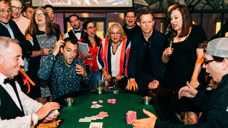 A group of people with shocked and happy expressions surround a green casino table and watch a card game unfold. 