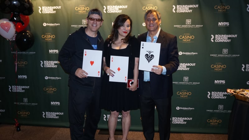 Two men and a woman stand in front of a green step and repeat with several logos. They each hold up a giant playing card, ace of hearts, diamonds, and spades.