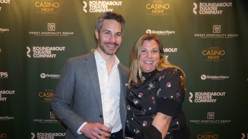 A man in a gray blazer with dark gray hair poses with a blonde woman wearing a black floral dress in front of a step and repeat.