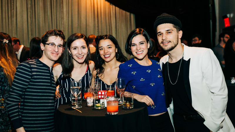 A group of five young adults gather around a black-clothed high top table decorated with drink glasses and smile for a photo.