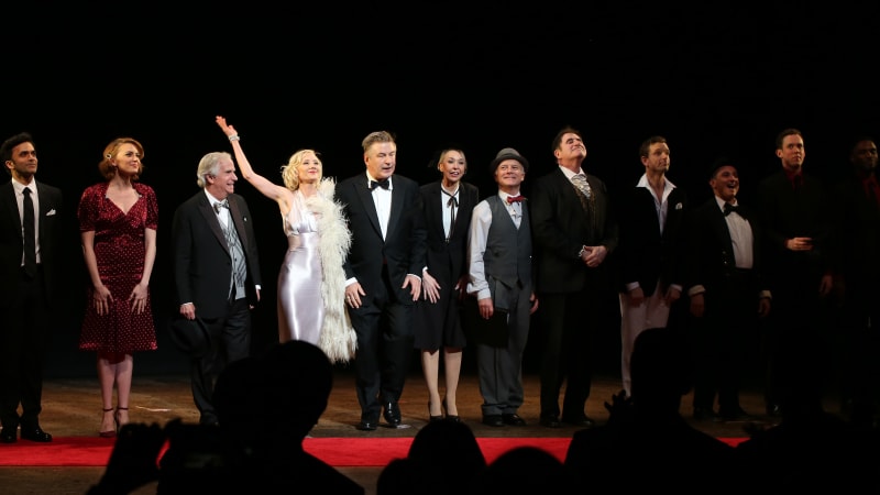 A line of actors on stage, wearing fancy suits and dresses, stand next to each other to take their bow.