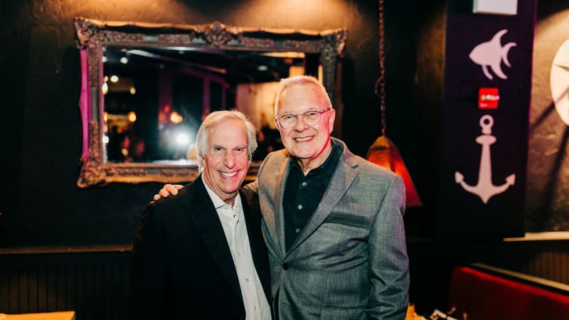 Two older men with white hair wear suits and pose for a photo with their arms around each other at a restaurant.