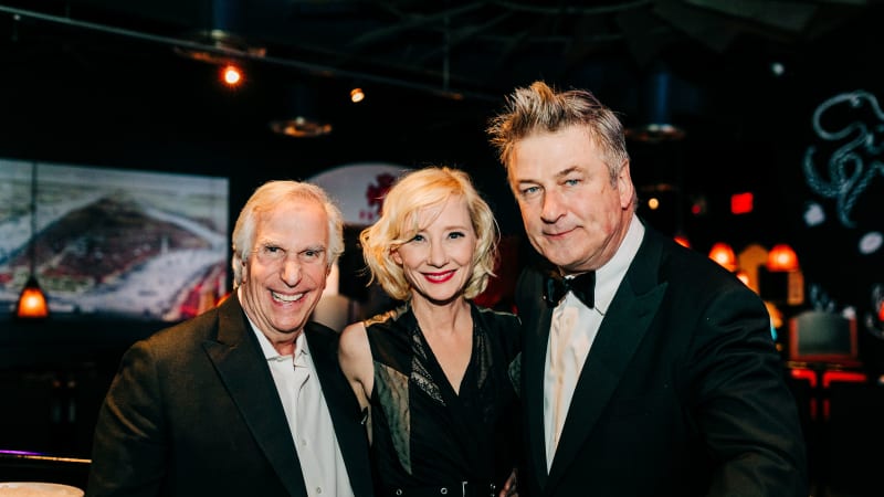 Two men in suits stand on either side of a woman with short curly blonde hair wearing a v-neck, belted black dress.