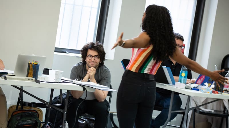 A man with long hair and round glasses smils while watching an actress sing with her arms outstretched in the rehearsal studio.