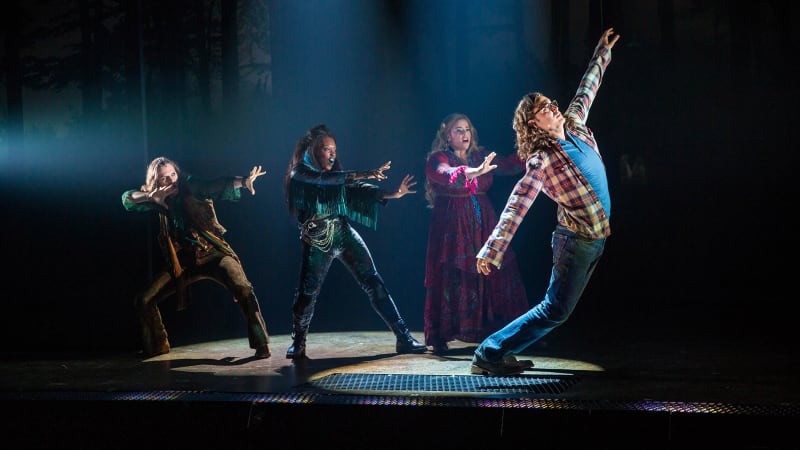 Three actors in bright, jewel-toned, bohemian outfits hold their arms like they're casting a spell. In front of them, a man in a flannel and glasses slowly falls to the floor.