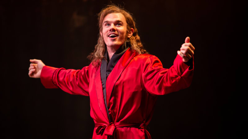 A man with long hair smiles and holds his arms outstretched. He wears a lavish, silk red robe.