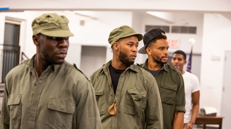 A line of Black actors dressed as soldiers. They look over their left shoulder with solemn looks.