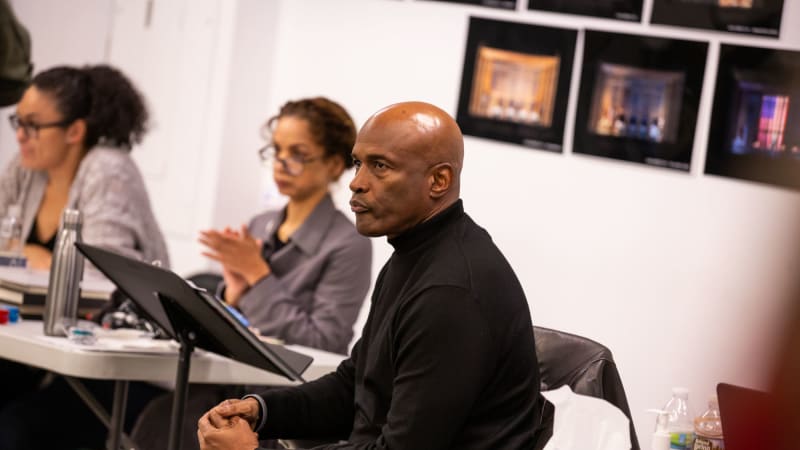 A Black bald man dressed all in black sits behind a music stand and watches rehearsal intently.