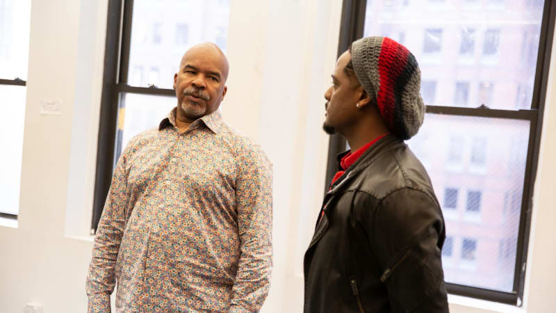Two Black men in the rehearsal studio, one holding his hands clasped behind his back like a soldier, the other looking at him with skepticism.