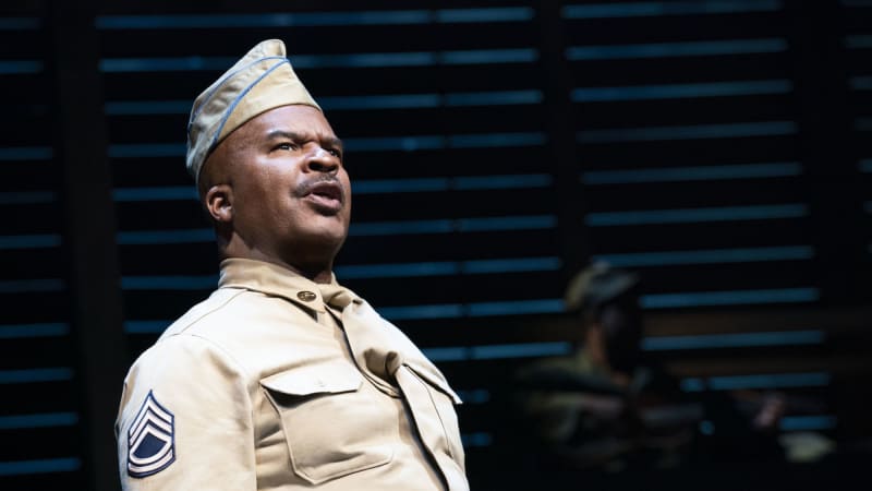 A Black man with a mustache wearsr a tan soldier's uniform and garrison cap. He speaks towards the audience with a determined expression.