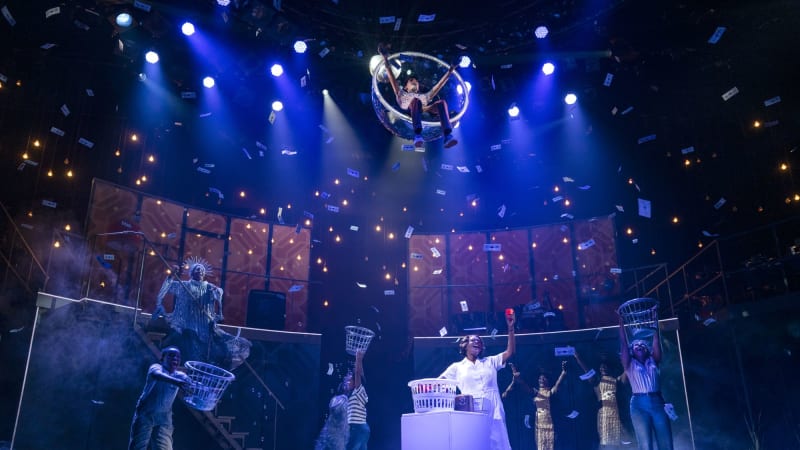 An elaborate musical number with blue lighting and dollar bills falling from the sky. Actors excitedly extend laundry baskets in the air. A Black woman raises a cup and sings by a laundry machine.