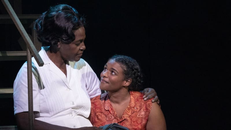 A Black woman in a white maid's dress sits on steps with her arm around a young, smiling Black woman wearing an orange paisley shirt and jeans.