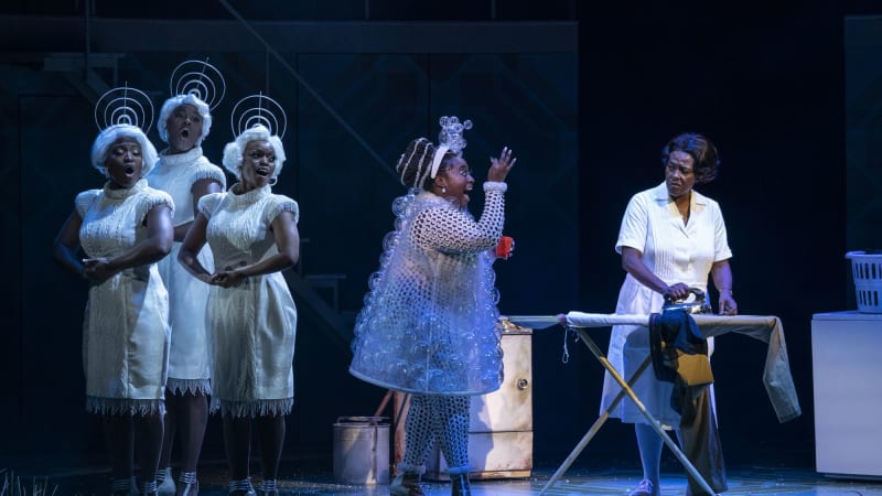 Three Black woman in white wigs and elaborate white costumes sing in a group while a Black woman in a costume made of bubbles sings by an unimpressed Black woman ironing a shirt