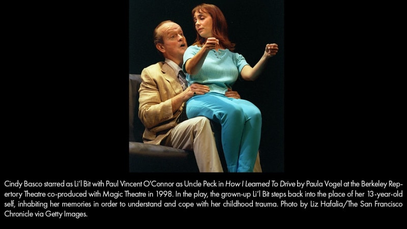 A light-skinned, brown-haired woman wearing a turquoise outfit sits on the lap of an older, light-skinned man in a tan suit. She holds an imaginary steering wheel