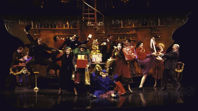 The company of She Loves me all in jackets and hats on stage. The set is of a bar and everyone is celebrating with gifts.
