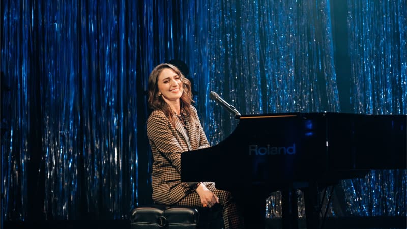 Sara Barellies, a woman with light skin and wavy brown hair, wears a checkered suit and sits at a black piano on stage in front of a blue sparkly backdrop.