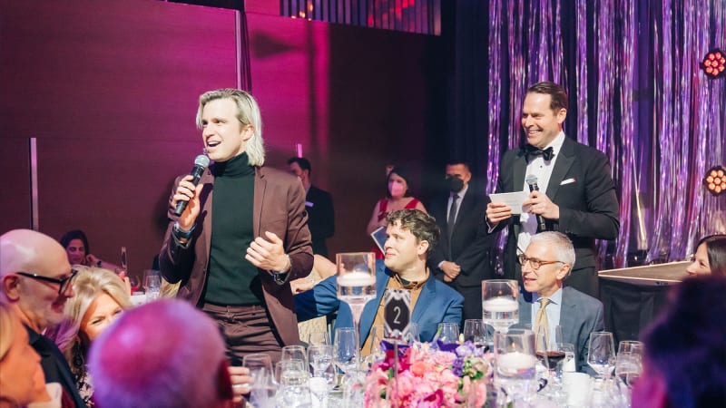 Two people stand and hold micrphones in a room full of people sitting at fancy dining tables under a wash of pink and purple lighting.