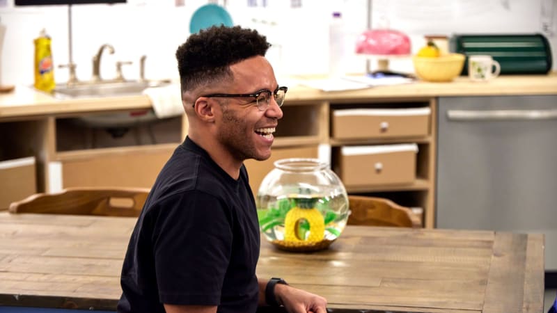 A Black man wearing glasses laughs while sitting at a wooden table in a rehearsal set that looks like a kitchen. 