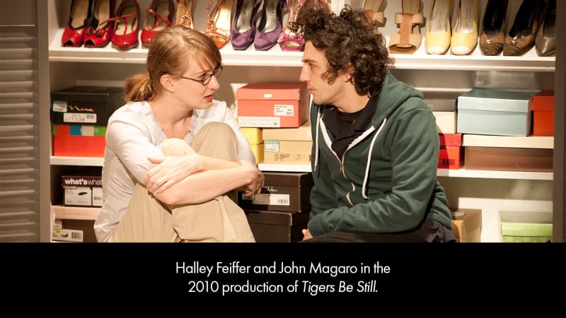 Production image from TIGERS BE STILL. A teenage girl wearing tan pants and a blue sweater and a teenage boy wearing a green hoodie sit on the floor of a shoe closet.