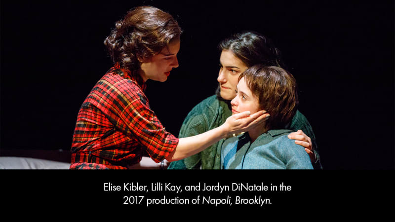 Production photo from NAPOLI, BROOKLYN. Three light-skinned, dark-haired young women in 1960s clothing sit together on a bed.