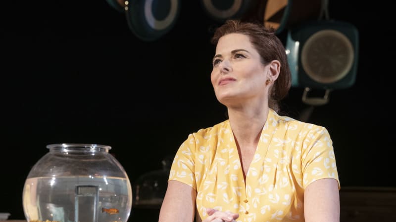 A woman with auburn hair wears a yellow dress and sits at a table with a fishbowl, in it a swimming goldfish.