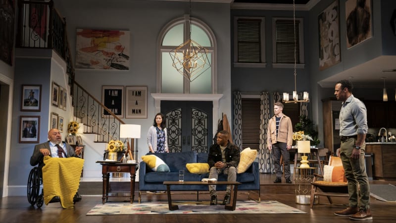 Five actors stand and sit around a set that looks like the living room of a wealthy home. The oldest actor sits in a wheelchair with a blanket on his lap and speaks to the room.