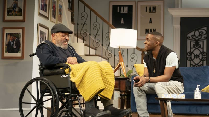 An old man in a wheelchair smiles and talks to a young man who sits close to him on a couch in a living room.