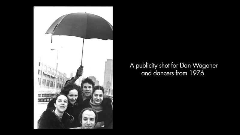 A publicity shot for Dan Wagoner and dancers from 1976. Five light-skinned adults stand under an umbrella held by a man at the back. They stand on a city rooftop.