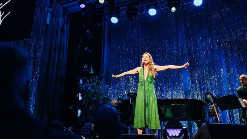 Sutton Foster sings on stage in in a sparkly green dress in front of a blue sparkly curtain.