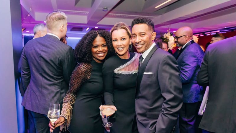 Three people in fancy attire pose together and smile while holding beverages.