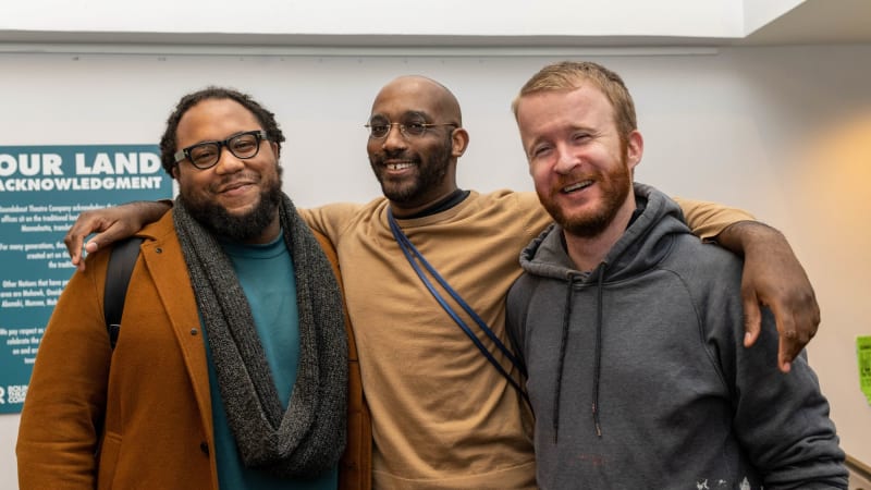 Three people smile and laugh together for a photo while the middle person puts their arms around the other two.