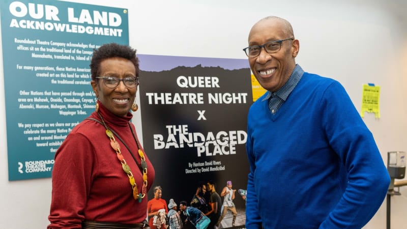 Two older people smile and stand on either side of an easel with a sign on it that says QUEER THEATRE NIGHT X THE BANDAGED PLACE.