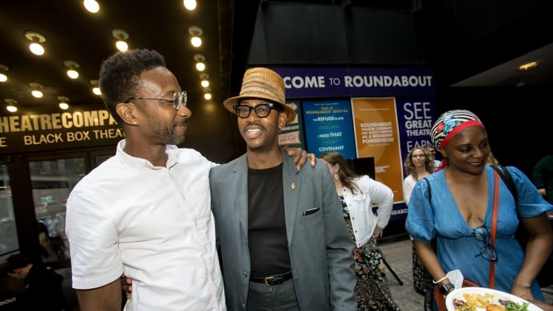 A man in a white button up shirt and classes puts his arm around Eric Berryman, who wears a straw, fedora hat, black glasses, and a blazer.