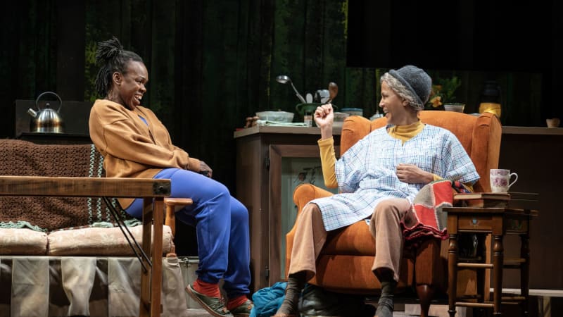 A young Black woman sits across from and laughs with an old Black woman sitting in an orange recliner chair.