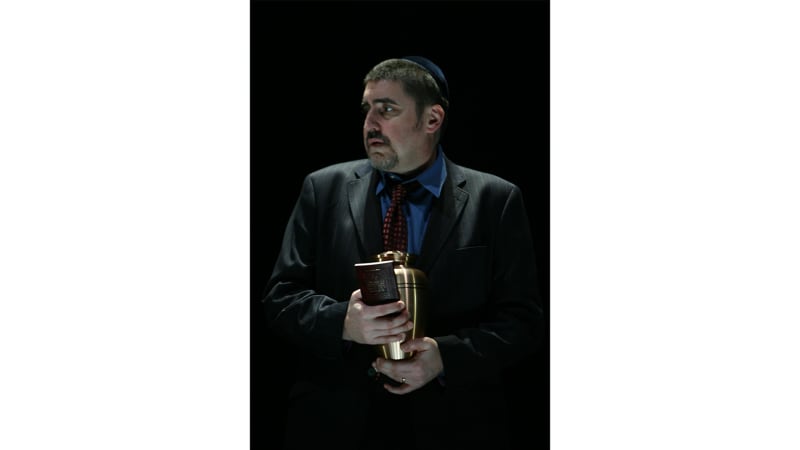 A light-skinned man with dark hair and a goatee wears a dark suit and yarmulke. He holds an urn and a prayer book.
